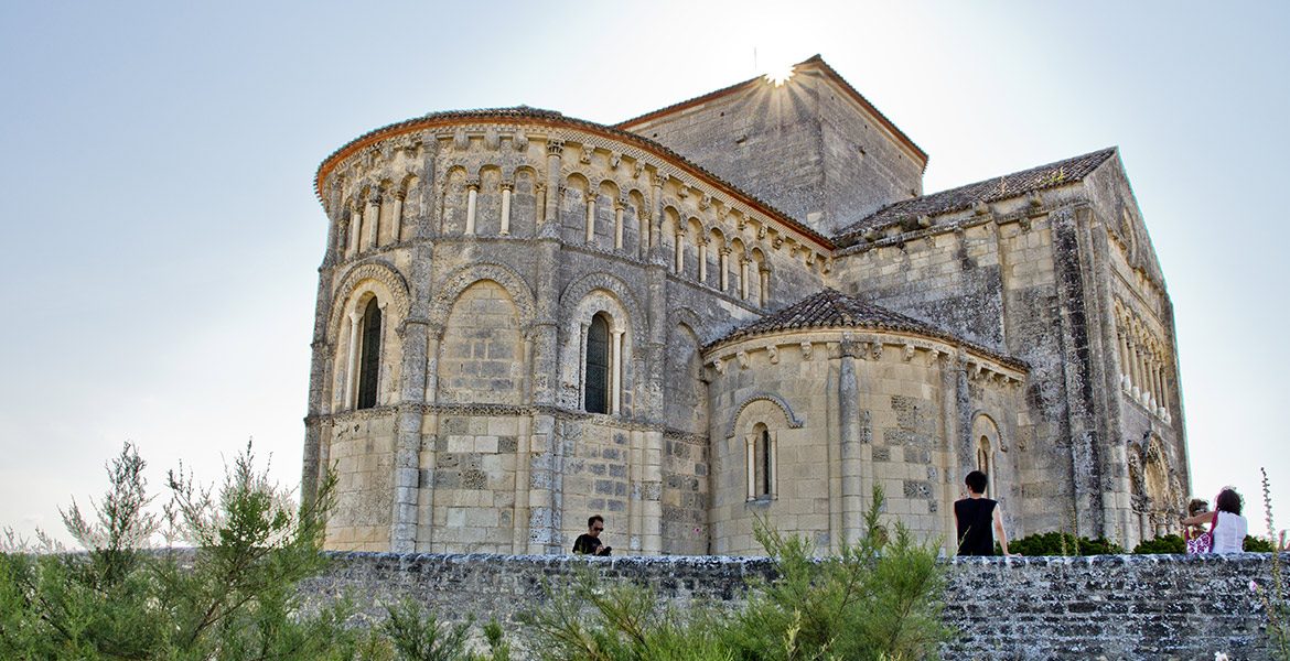 Vue de l'église Sainte-Radegonde de Talmont-sur-Gironde, en Charente-Maritime, avec ses arcs romans et sa pierre calcaire. Des visiteurs se promènent autour, admirant l'architecture historique sous un ciel clair.