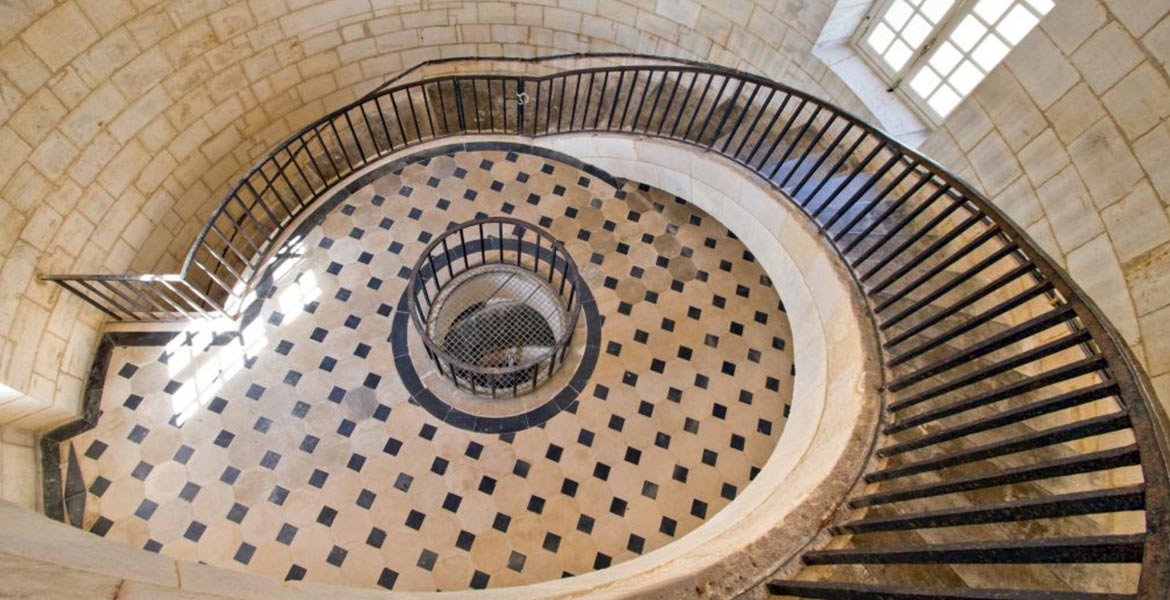 Vue aérienne de l'escalier en colimaçon du Phare de Cordouan avec un sol carrelé noir et blanc. La lumière naturelle entre par une fenêtre, éclairant l'élégante architecture intérieure.