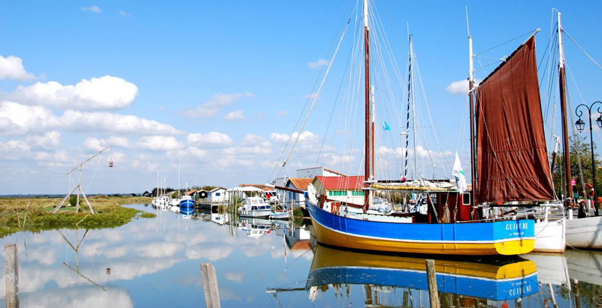 Bateau de pêche coloré amarré dans le port de Mornac sur Seudre, avec des maisons et d'autres bateaux en arrière-plan sous un ciel bleu parsemé de quelques nuages blancs.