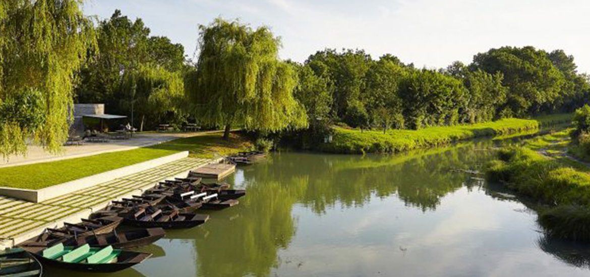 Des barques noires sont amarrées le long d'un quai en bordure d'une rivière entourée de verdure luxuriante et de saules pleureurs. L'endroit est paisible, propice à la détente et à la promenade en pleine nature.