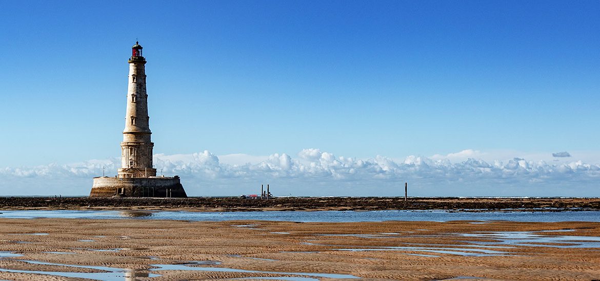 Phare de Cordouan avec des motifs de marée basse, entouré par l'océan sous un ciel bleu clair avec des nuages à l'horizon.