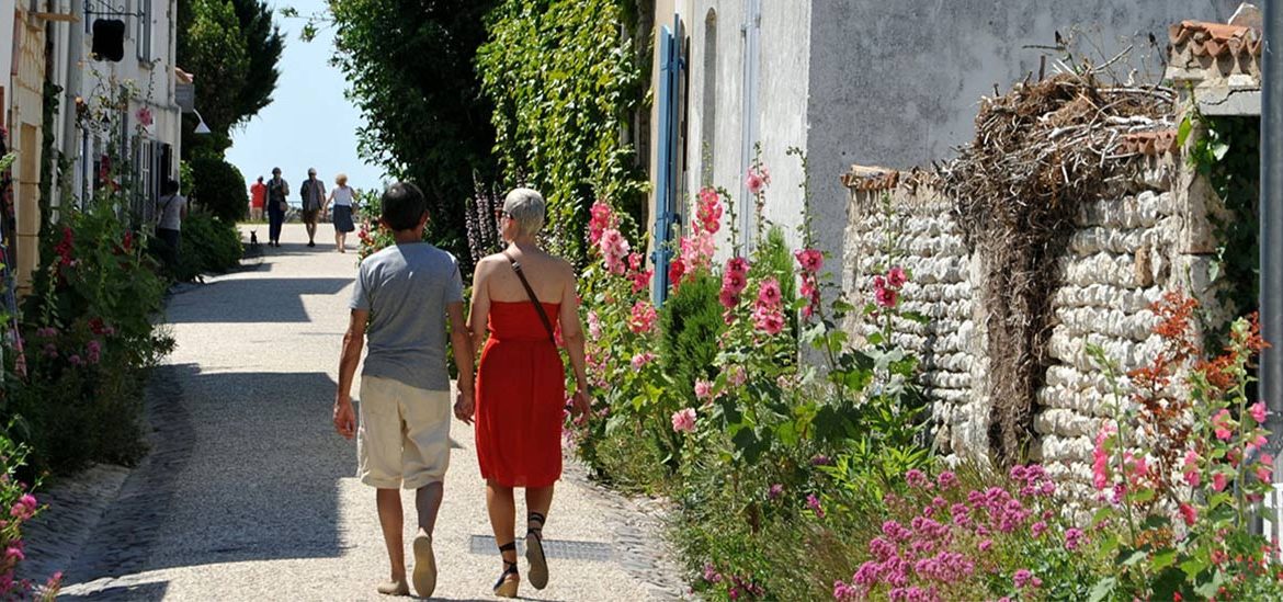 Un couple se promène main dans la main dans une ruelle fleurie de Talmont-sur-Gironde, en Charente-Maritime, bordée de maisons traditionnelles aux volets bleus. La scène est paisible et baignée de lumière estivale.