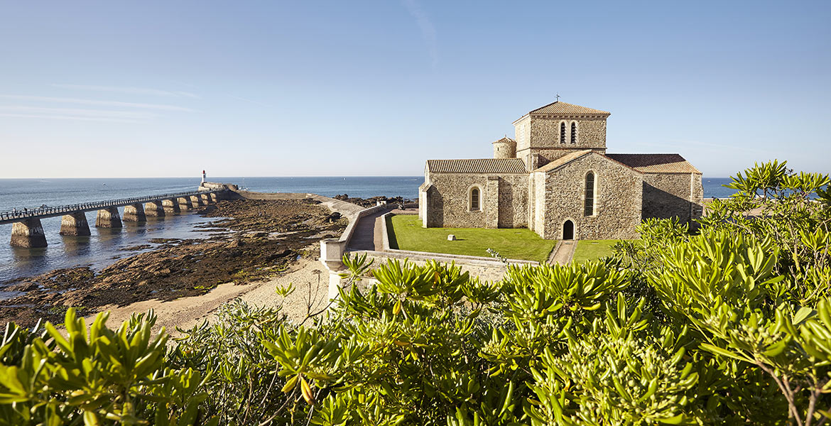 Vue de l'église Saint-Nicolas de la Chaume et de la jetée Jacobsen à Les Sables-d'Olonne, surplombant l'océan Atlantique. Le ciel est dégagé, offrant un paysage pittoresque et paisible.