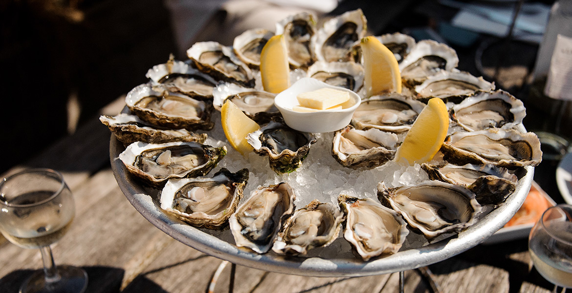 Un plateau d'huîtres fraîches disposées sur de la glace, accompagné de quartiers de citron et d'un petit bol de beurre. Une assiette et un verre de vin blanc sont placés à côté sur une table en bois.