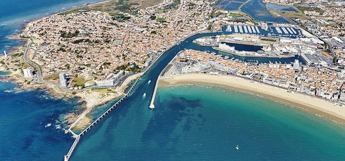 Vue aérienne des Sables-d'Olonne, montrant la plage de sable doré, le port de plaisance et les jetées s'avançant dans l'océan. Les toits rouges des bâtiments et les eaux turquoises créent un paysage côtier pittoresque.