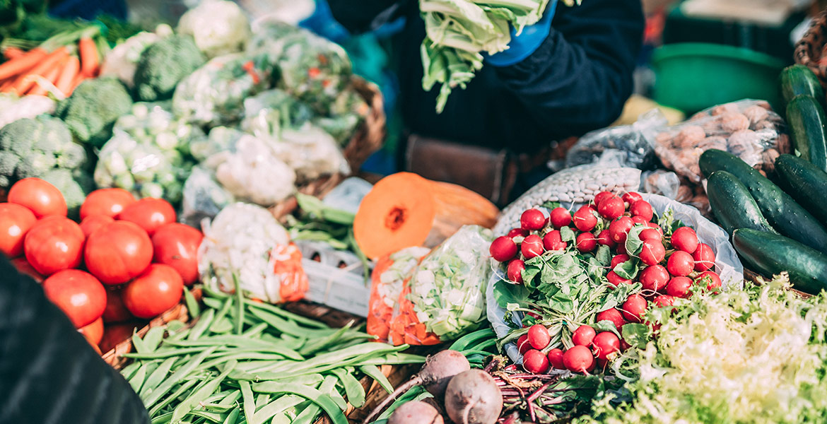 Un étal de marché rempli de légumes frais, incluant des tomates, des radis, des haricots verts, des concombres, des carottes, et des brocolis. Une personne en arrière-plan manipule des légumes verts avec des gants bleus.