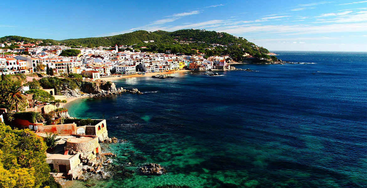 Vue panoramique de la ville côtière de Calella de Palafrugell, avec ses maisons blanches et colorées bordant la mer turquoise. Les collines verdoyantes et le ciel dégagé complètent ce paysage pittoresque de la Costa Brava.