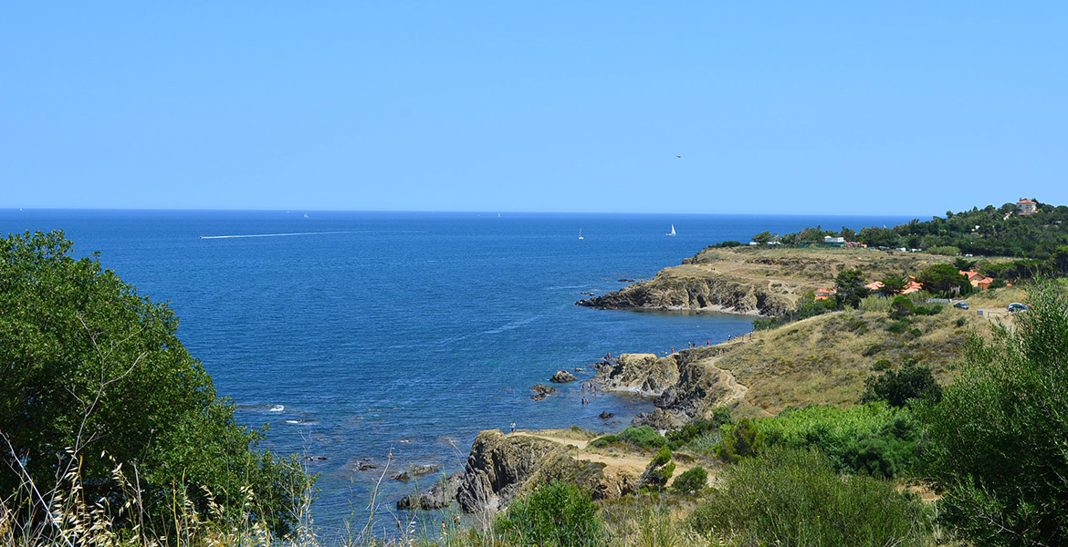 Vue panoramique d'une côte rocheuse avec des sentiers de randonnée, des baigneurs et des voiliers sur une mer calme. La végétation méditerranéenne entoure la scène sous un ciel bleu éclatant.