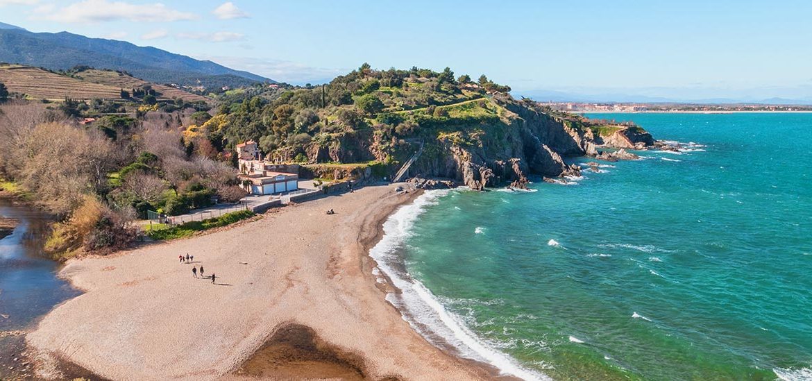 Vue aérienne d'une plage de galets avec des promeneurs le long du rivage et des collines verdoyantes en arrière-plan. La mer turquoise s'étend à perte de vue sous un ciel dégagé.