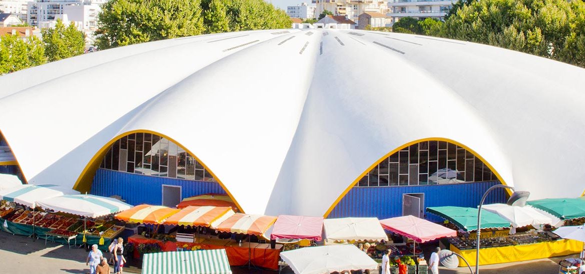 Le marché central de Royan animé avec des stands colorés se trouve devant un bâtiment en forme de coquillage. Des personnes se promènent et font leurs courses parmi les étals de fruits et légumes.