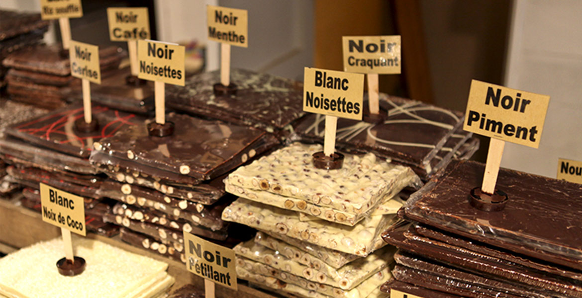 Tablette de chocolats variés avec des étiquettes indiquant les saveurs telles que "Noir Noisettes" et "Blanc Noix de Coco". Les tablettes sont empilées sur un stand de marché.