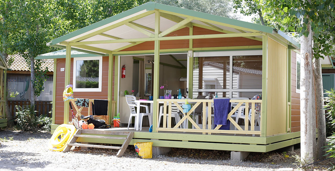 Un chalet de camping en bois avec une terrasse, entouré d'arbres et équipé de jouets et de chaises.