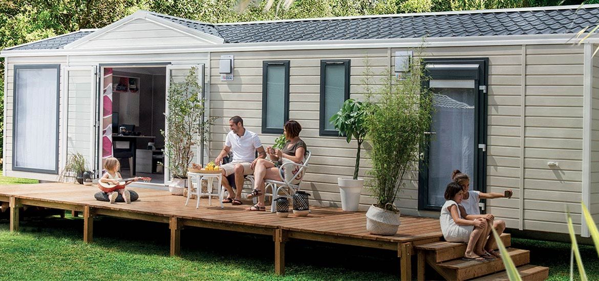 Une famille profite du soleil sur la terrasse en bois d'un mobil-home. Un enfant joue de la guitare, tandis que les parents discutent autour d'une table avec des fruits. Deux autres enfants sont assis sur les marches. Des plantes en pot décorent la terrasse.