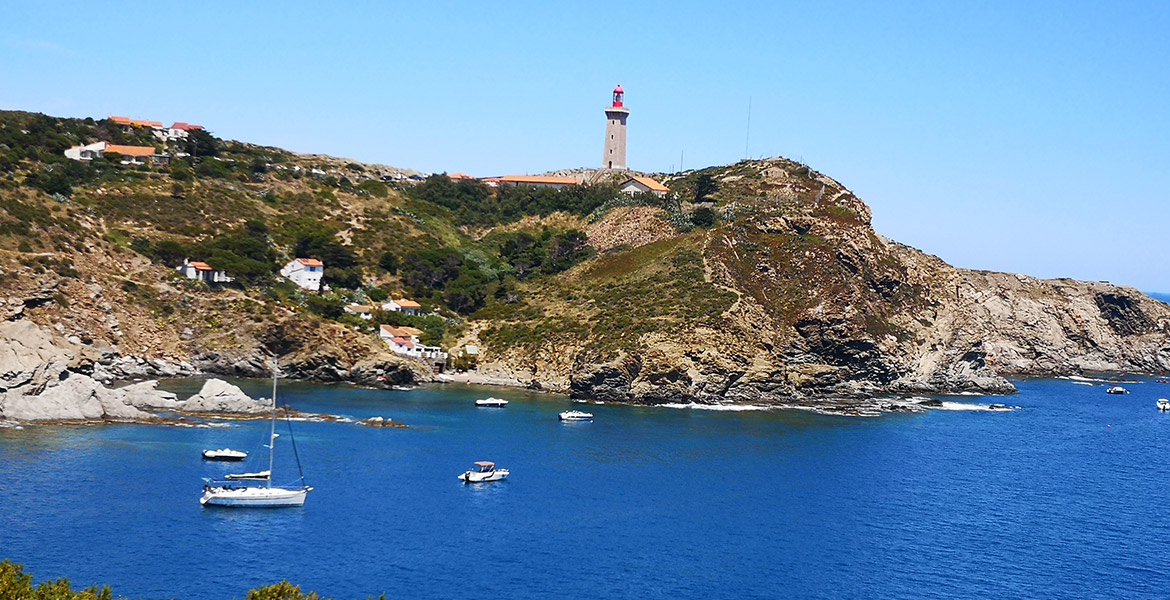 Des bateaux sont ancrés dans une baie près d'un phare sur une colline rocheuse. La mer bleue et le paysage côtier créent une scène pittoresque et tranquille.