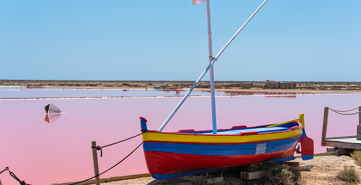 Un bateau de pêche coloré est amarré sur la rive d'un marais salant aux eaux roses sous un ciel bleu.