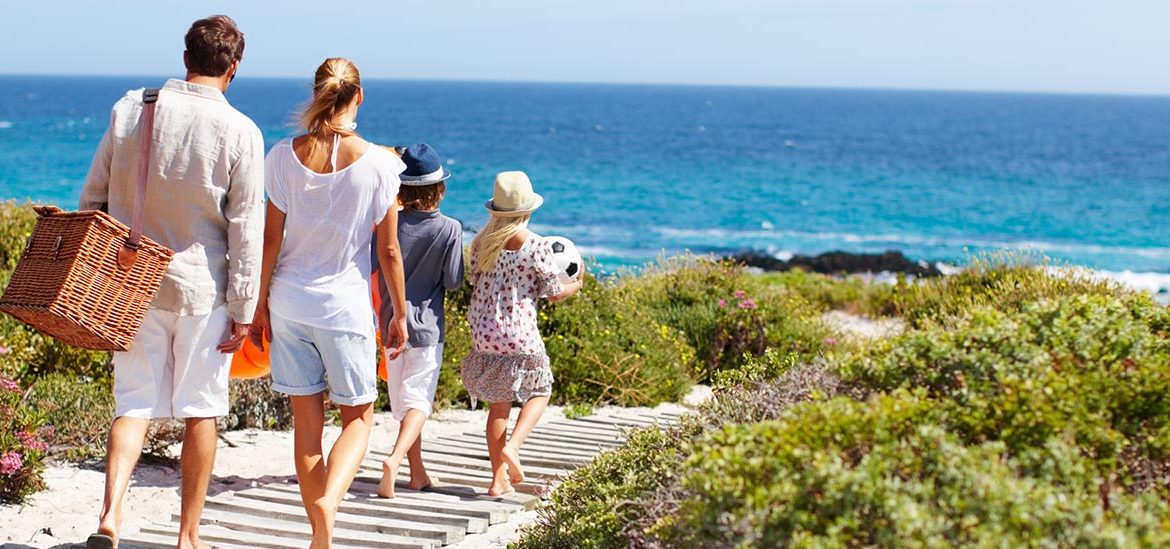 Une famille marchant vers la plage, portant un panier de pique-nique et un ballon, avec la mer en arrière-plan.