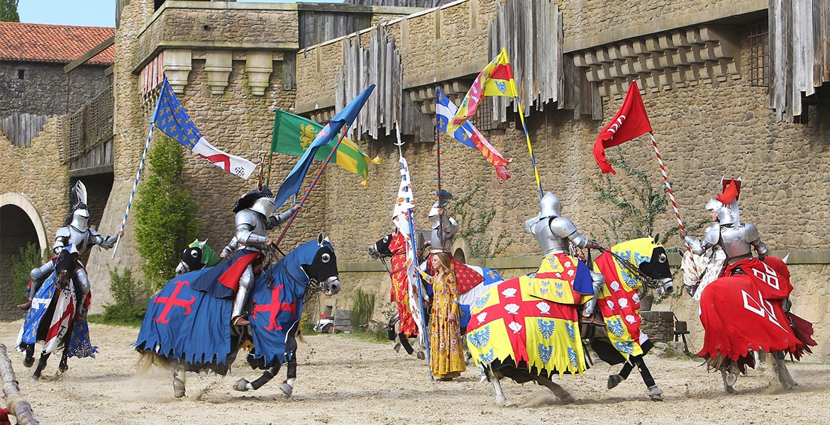 Des chevaliers du Puy du Fou en armure montent des chevaux décorés de caparaçons colorés, participant à un tournoi médiéval devant un château en pierre.