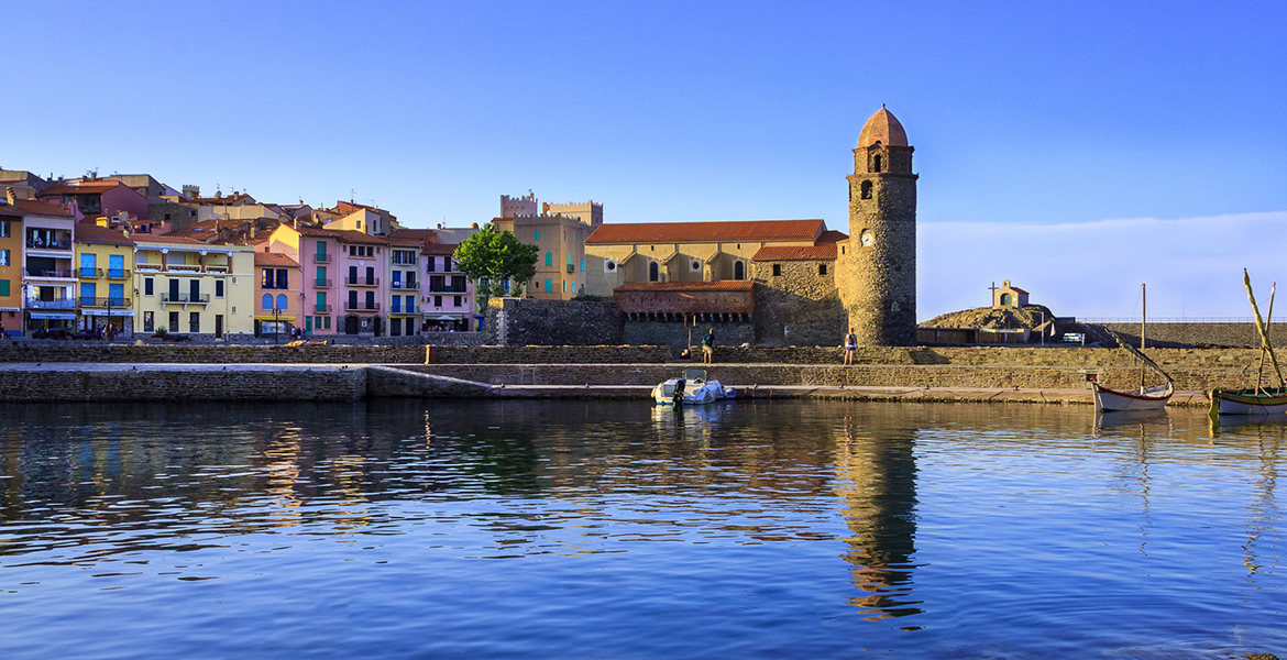 Un village côtier avec des maisons colorées, une église en pierre avec un clocher, et des bateaux amarrés dans l'eau calme sous un ciel bleu.