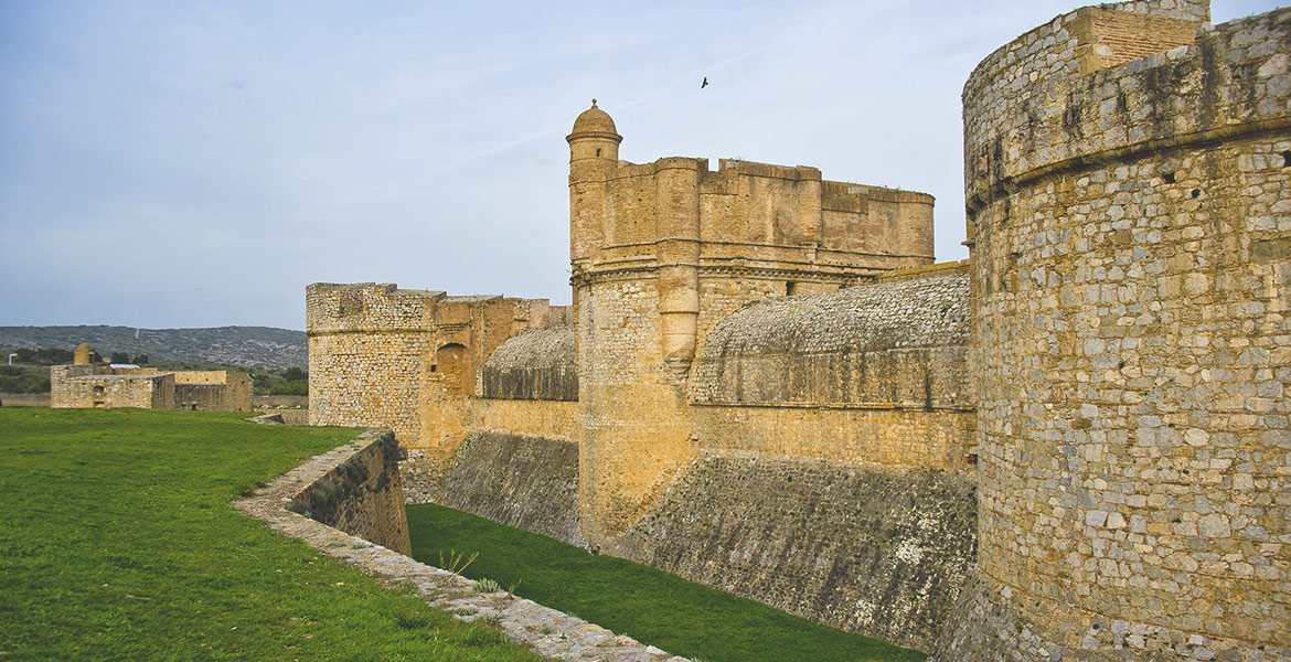 Les fortifications massives de la Forteresse de Sales en pierre, entourées de douves verdoyantes, avec un paysage de collines en arrière-plan.