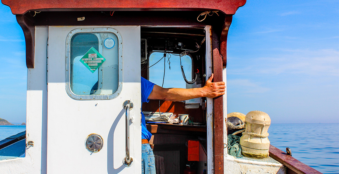 Un marin tenant la porte de la cabine d'un bateau, avec l'océan et un ciel bleu en arrière-plan.