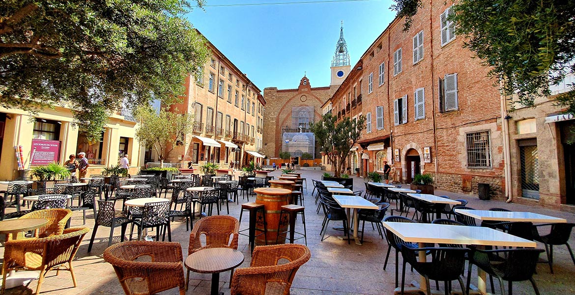 Une place ensoleillée de Perpignan avec des tables et des chaises de café en plein air, entourée de bâtiments historiques en briques, avec une église en arrière-plan.