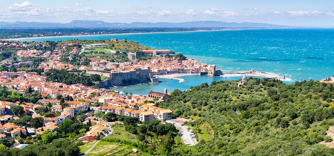 Vue panoramique sur un village côtier dans les Pyrénées Orientales avec des maisons aux toits rouges entourées de végétation verdoyante, donnant sur la mer turquoise.