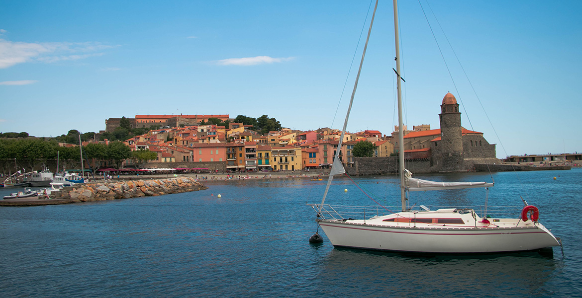 Un voilier blanc est amarré dans le port de Collioure, avec ses maisons colorées et son église en arrière-plan. Le ciel est dégagé, mettant en valeur le charme méditerranéen de la ville.