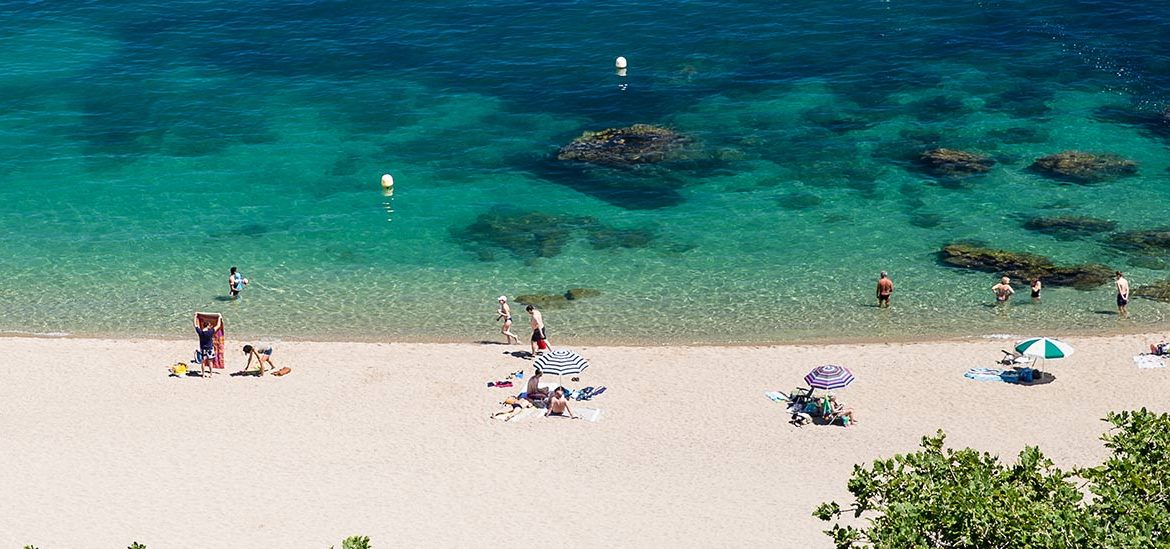 Des vacanciers profitent d'une plage de sable fin avec des parasols colorés, bordée par une mer turquoise et des rochers sous l'eau.