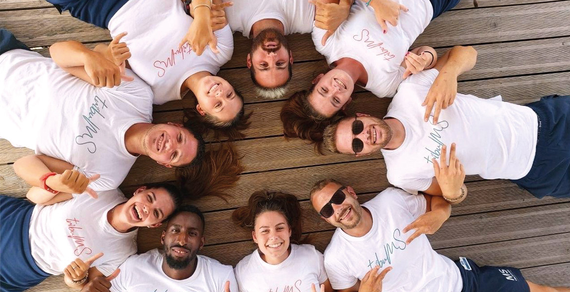 Groupe de neuf animateurs des Campings Campéole 4 étoiles et Camping Club MS 5 étoiles allongées en cercle sur un pont en bois, tous souriants et vêtus de t-shirts blancs et des Shorts avec inscrit dessus "Team ms"