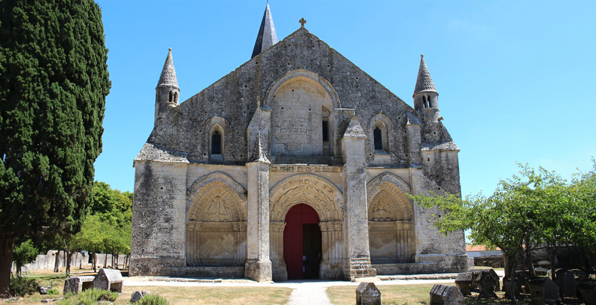 Façade d'une église en pierre avec une grande porte rouge et des détails architecturaux sculptés, entourée de verdure et de pierres tombales sous un ciel bleu.