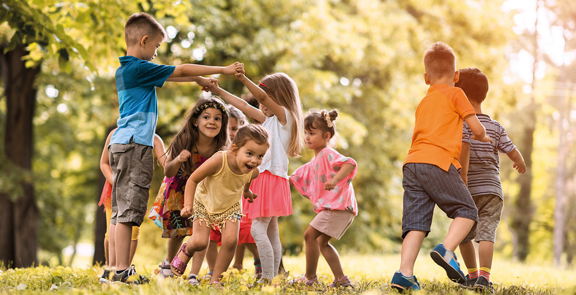 Un groupe d'enfants joue joyeusement en formant une ronde dans un parc verdoyant. Le soleil perce à travers les arbres, créant une atmosphère lumineuse et enjouée.