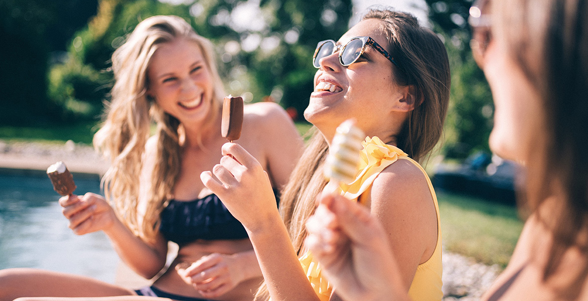 Trois jeunes femmes en maillot de bain rient ensemble en mangeant des glaces au bord d'une piscine. Le soleil éclatant et la verdure environnante créent une ambiance estivale joyeuse.