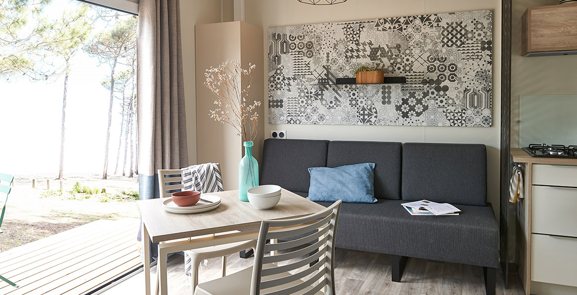 Intérieur lumineux et moderne d'un mobil-home avec un canapé gris, une table à manger décorée de vaisselle colorée et d'un vase, une grande fenêtre avec vue sur des arbres et de la nature à l'extérieur.