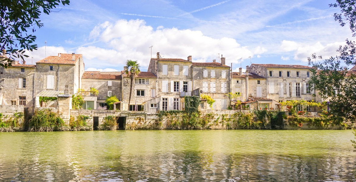 Maisons de Charente en pierre alignées le long d'une rivière, avec des balcons et des volets colorés, sous un ciel bleu avec des nuages blancs.