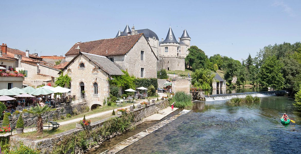 Une scène pittoresque d'un village avec des bâtiments en pierre, des terrasses de café au bord de l'eau, et un château en arrière-plan.