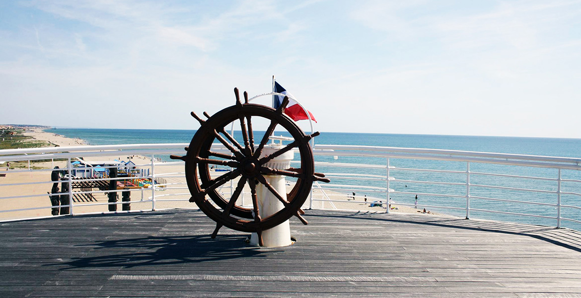 Une barre de navigation en bois sur le pont du paquebot "Lydia", avec le drapeau français en arrière-plan et une vue sur la plage et l'océan.