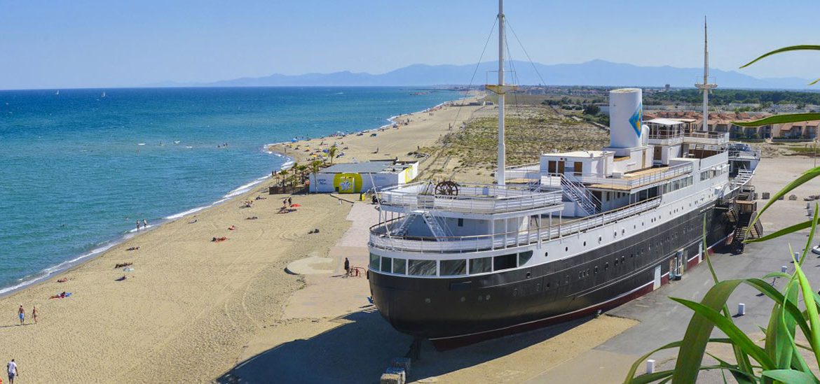 Le paquebot "Lydia" est échoué sur une plage de sable fin avec des baigneurs, l'océan et les montagnes en arrière-plan.