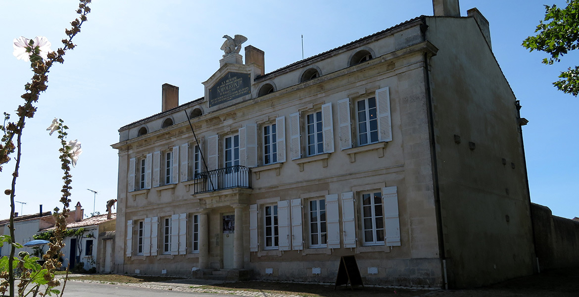 Musée napoléon III en Charente-Maritime en pierre avec des volets blancs, une plaque dédiée à Napoléon III sur la façade, sous un ciel bleu dégagé, avec des fleurs en avant-plan.