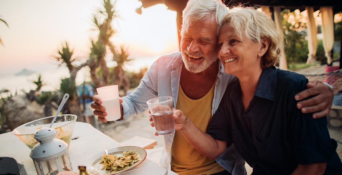 Un couple senior souriant partage un repas en plein air au coucher du soleil. Ils trinquent ensemble avec des verres d'eau, créant un moment de convivialité et de bonheur.