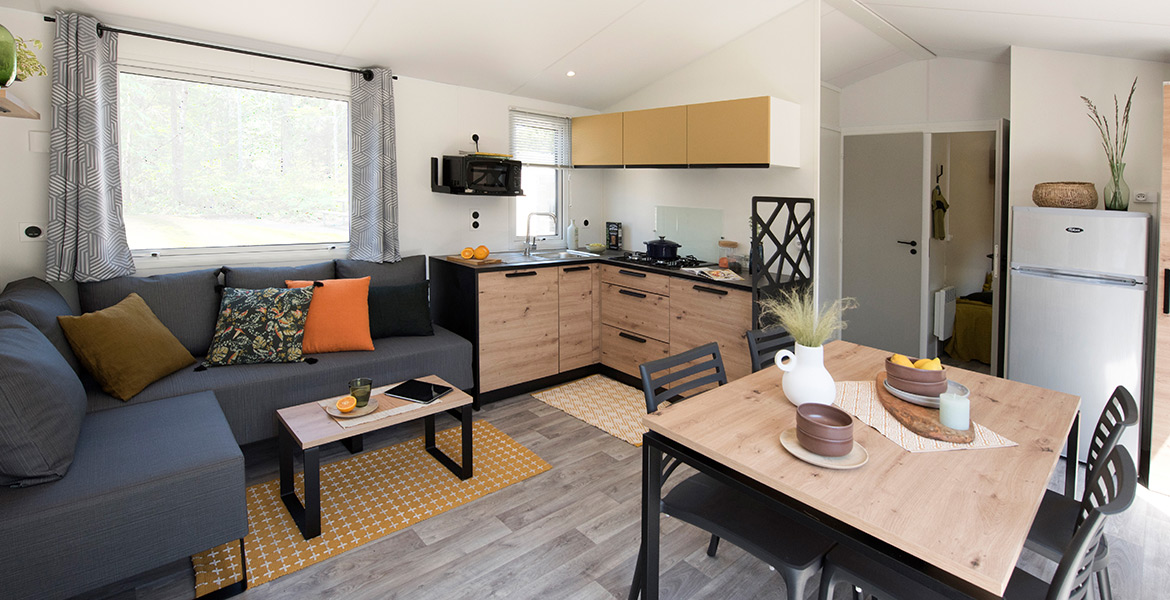 Intérieur moderne et lumineux d'un mobil-home avec un salon équipé d'un canapé gris, une cuisine avec des meubles en bois clair, et une table à manger décorée, sous une grande fenêtre donnant sur l'extérieur verdoyant.