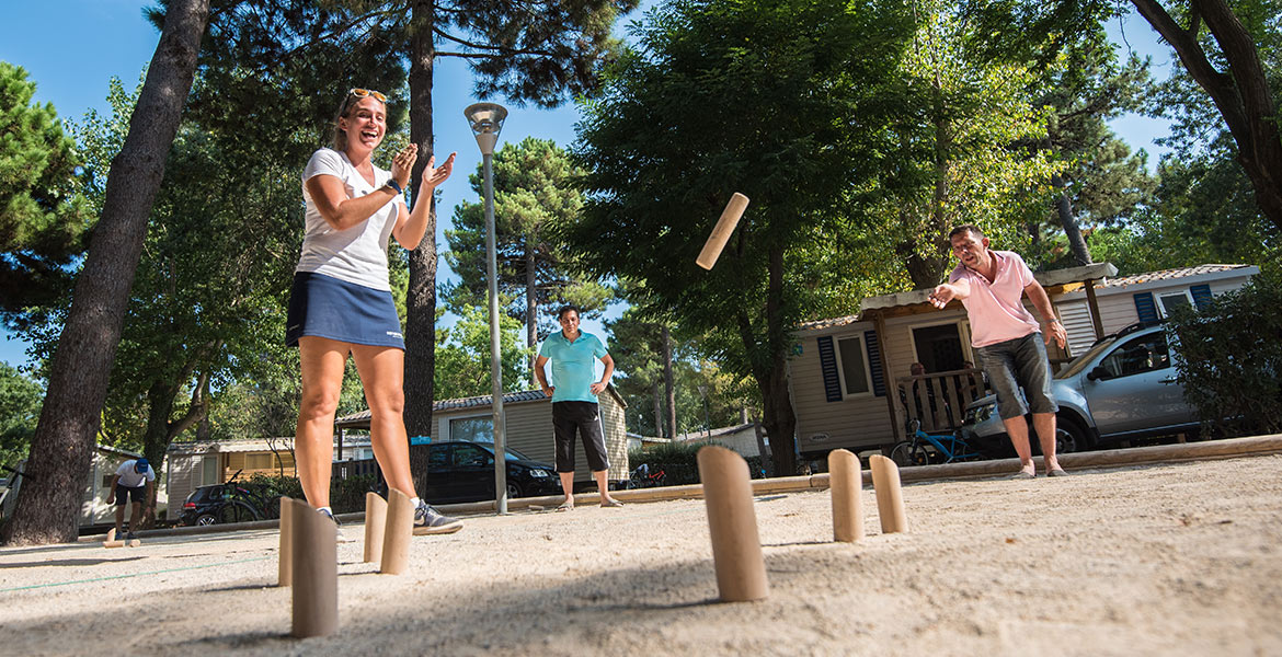 Trois personnes jouant à un jeu de quilles finlandaises (Mölkky) en extérieur dans un camping, entourées d'arbres et de mobil-homes, sous un ciel ensoleillé.