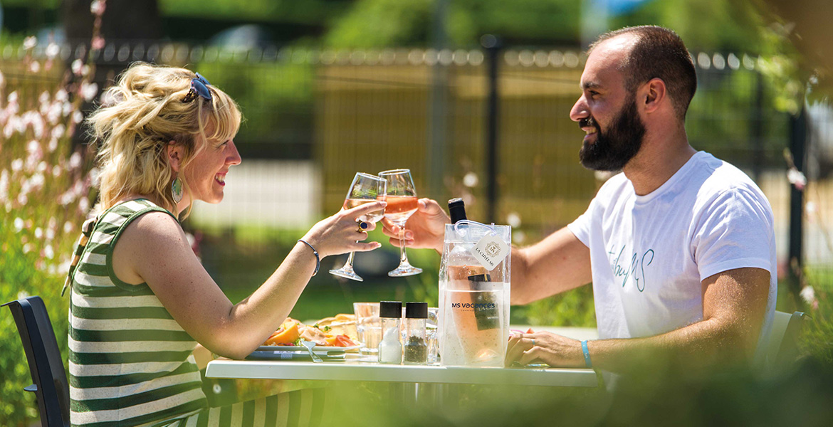 Un homme et une femme trinquent avec des verres de vin rosé, assis à une table en plein air, souriants et entourés de verdure par une journée ensoleillée.