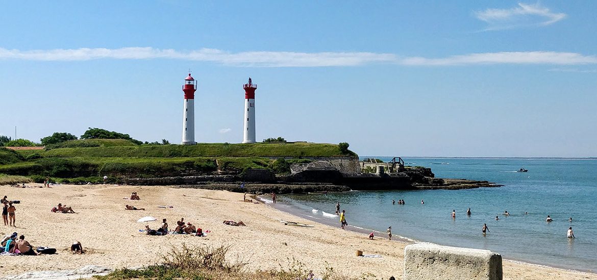 Plage à l'Île d'Aix avec des baigneurs et des promeneurs, surplombée par deux phares rouges et blancs situés sur une colline verdoyante sous un ciel bleu dégagé.