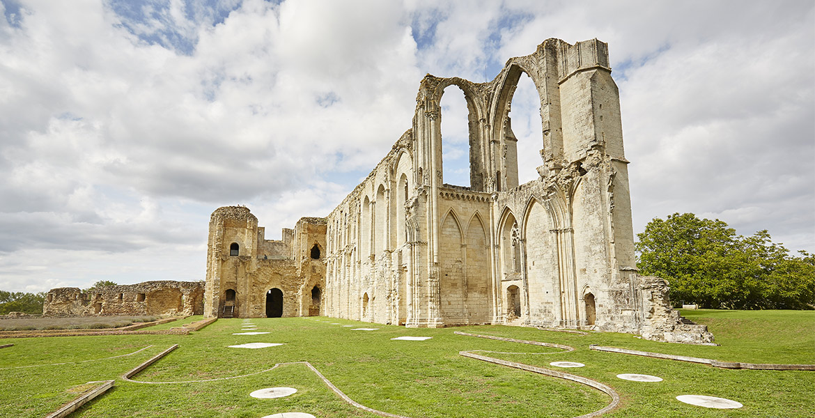 Ruines de l'Abbaye médiévale de Maillezais en Vendée, avec des arches imposantes, entourées d'une pelouse verte et sous un ciel partiellement nuageux.