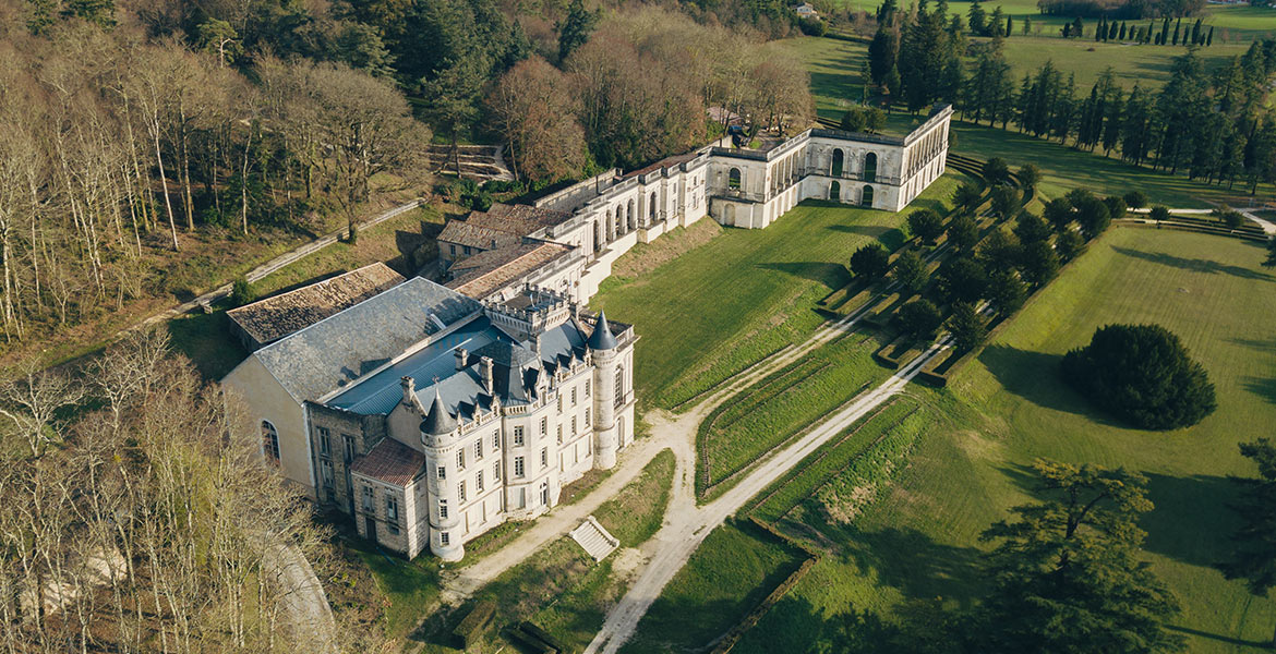 Vue aérienne du Château de la Mercerie entouré de vastes jardins et d'arbres, avec des allées bien entretenues et des bâtiments annexes.