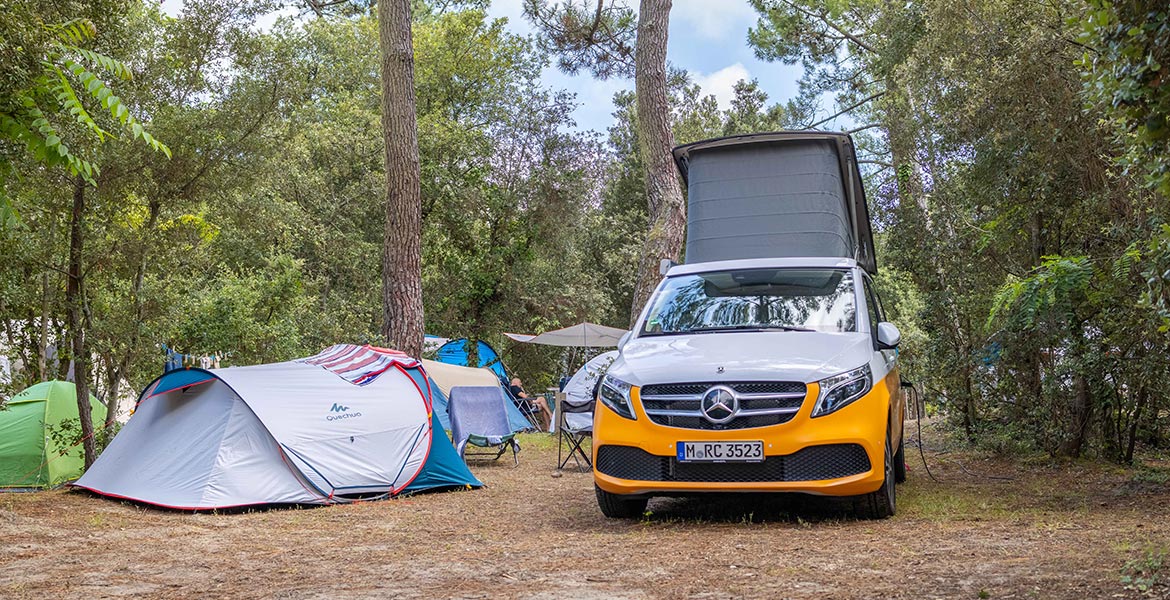 Camping en forêt avec une tente Quechua blanche et bleue à côté d'un van Mercedes-Benz orange et blanc. Des arbres entourent le site, créant une atmosphère naturelle et ombragée.