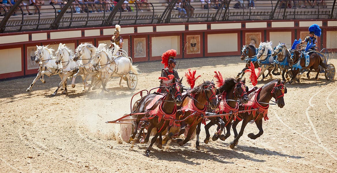 Course de chars romains dans une arène, avec des chevaux noirs en armure rouge et des chevaux blancs en armure bleue, dirigés par des conducteurs en tenue historique au Puy du Fou en Vendée.