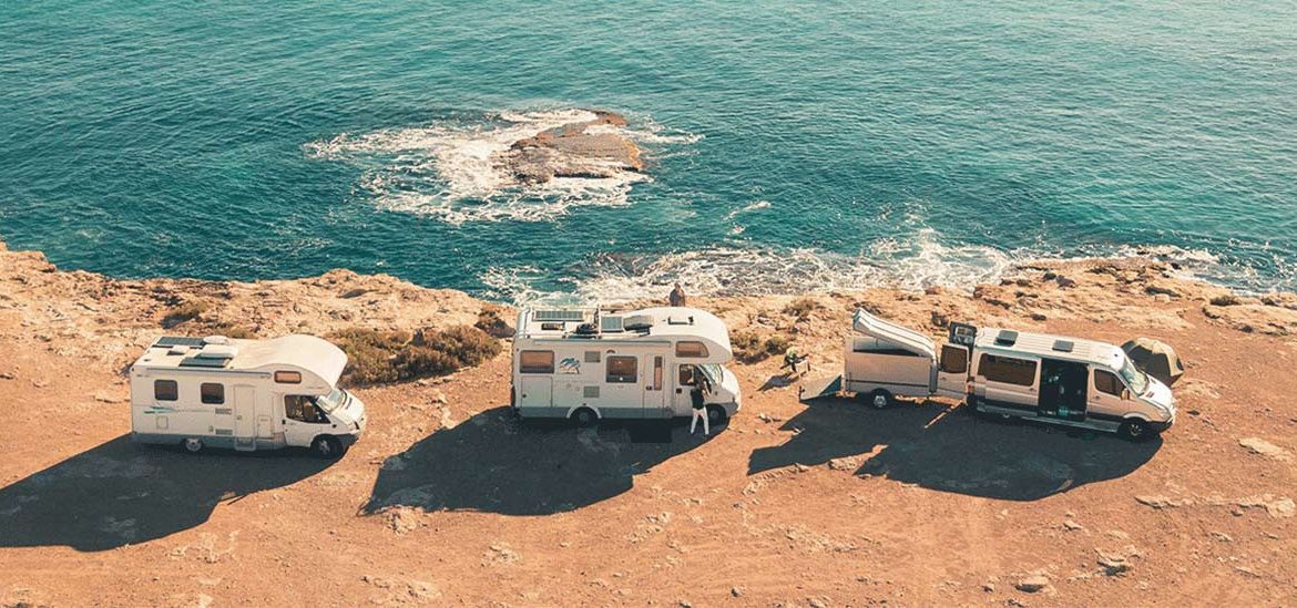 Trois camping-cars stationnés au bord d'une falaise rocheuse surplombant une mer bleue avec des vagues se brisant sur les rochers.