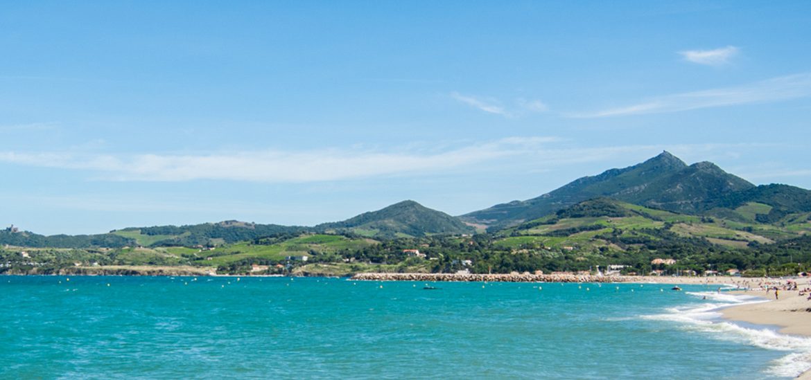 Plage de sable avec des vagues, bordée de collines verdoyantes sous un ciel bleu clair dans les Pyrénées Orientales.