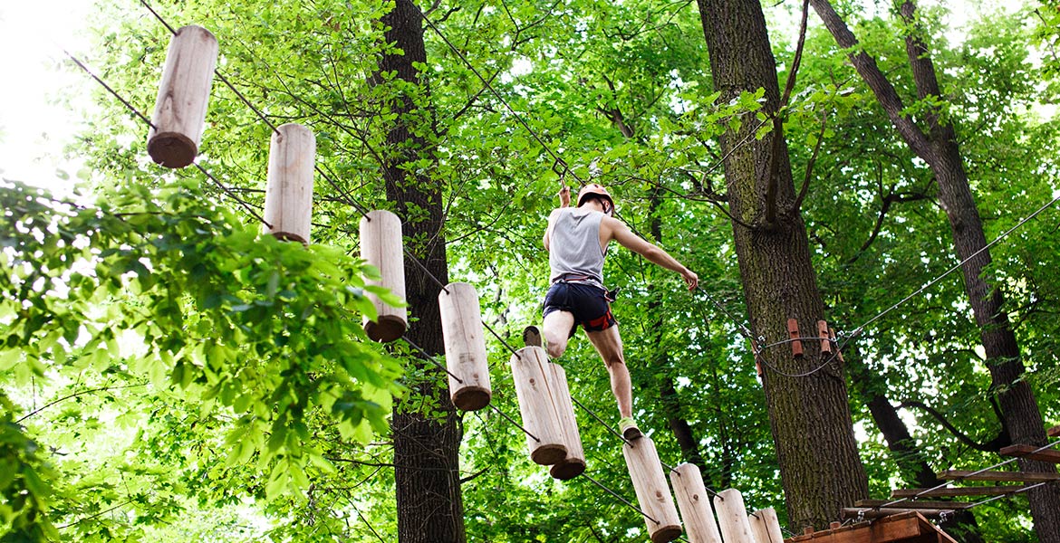 Une personne traversant un parcours d'accrobranche dans une forêt, marchant sur des rondins de bois suspendus, équipée d'un casque de sécurité et d'un harnais.